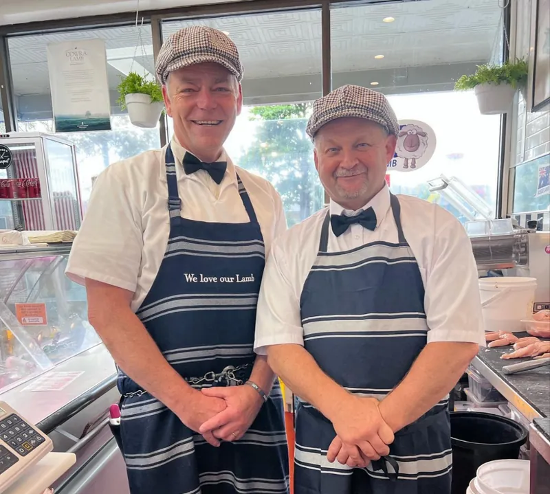 Two of the butchers behind the counter in houndstooth flat caps, bow ties and striped aprons, one reading 'We love our Lamb'